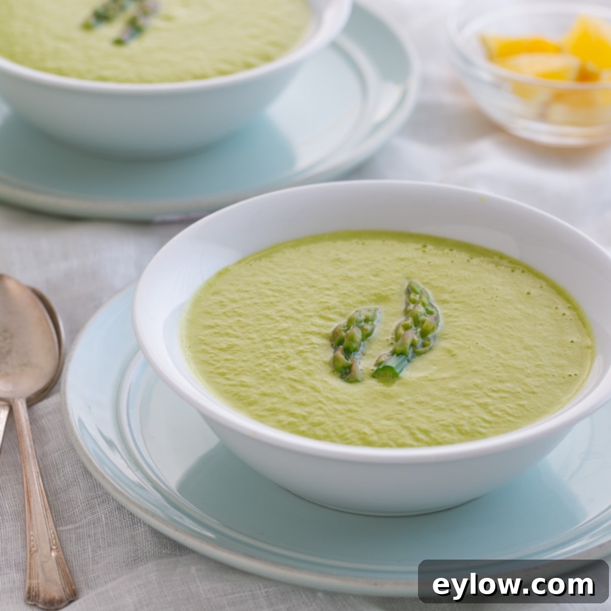 Bright green asparagus soup in a white bowl on a blue plate, set on a dinner table ready for serving.