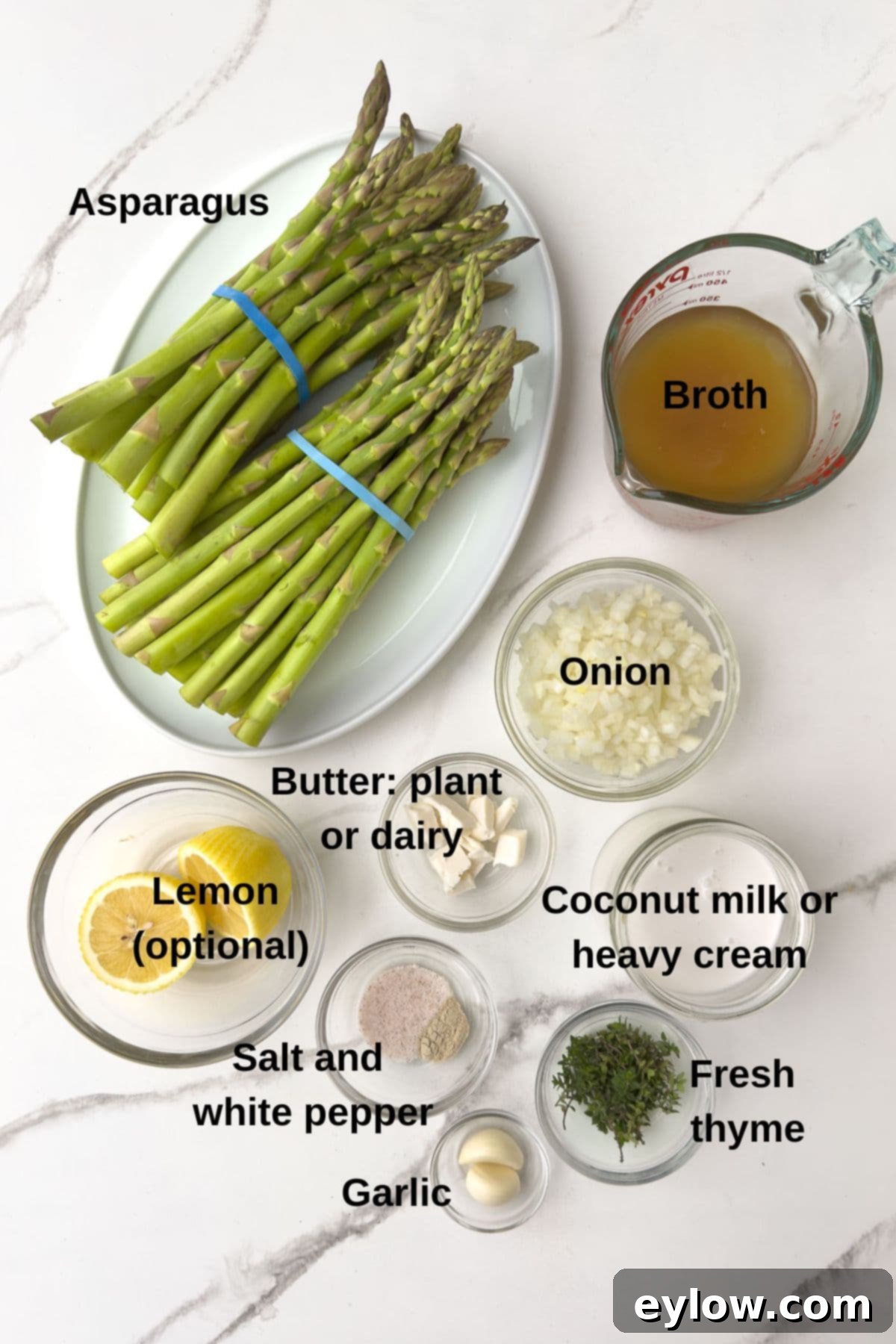 Ingredients for creamy asparagus soup laid out on a counter in prep bowls, including fresh asparagus, onion, garlic, broth, and cream options.