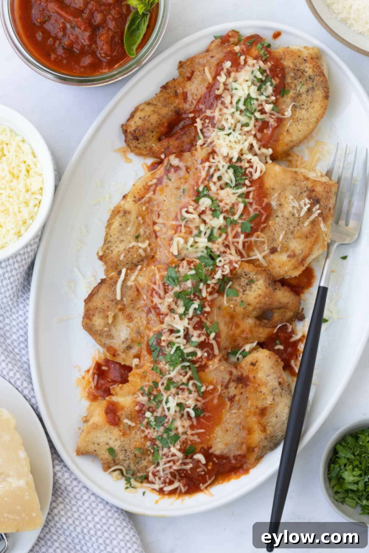A white platter of crispy chicken parmesan with red sauce and sprinkles of cheese, garnished with fresh basil.