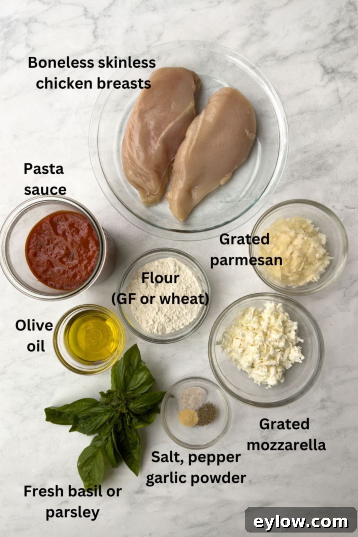 Ingredients for chicken parmesan laid out on a kitchen counter, including chicken breasts, cheese, sauce, and herbs.