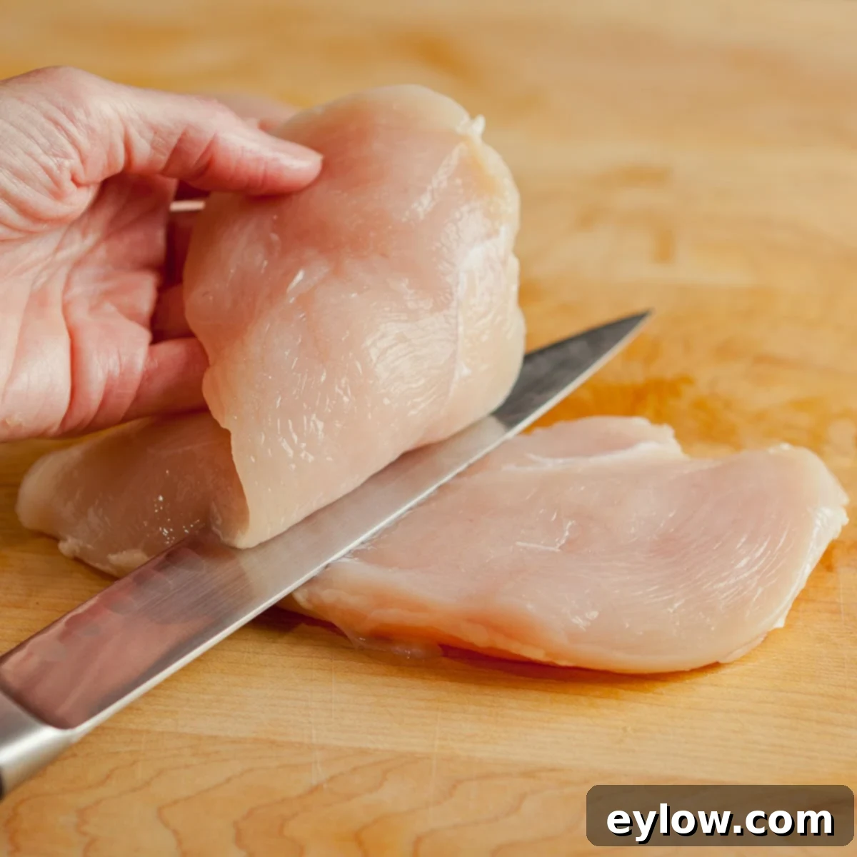 A chicken breast being precisely sliced into half for a cutlet with a thin sharp knife, parallel to the cutting board.