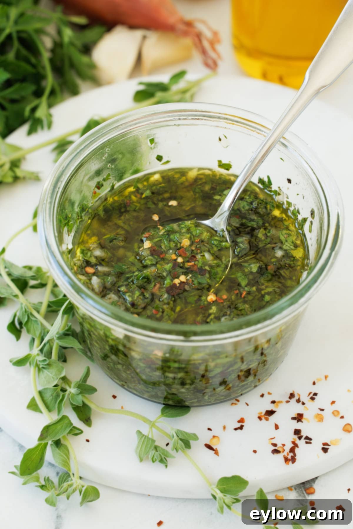 Authentic Argentinian Chimichurri Sauce 2 A jar of bright green herb sauce called chimichurri, surrounded by fresh parsley and oregano.