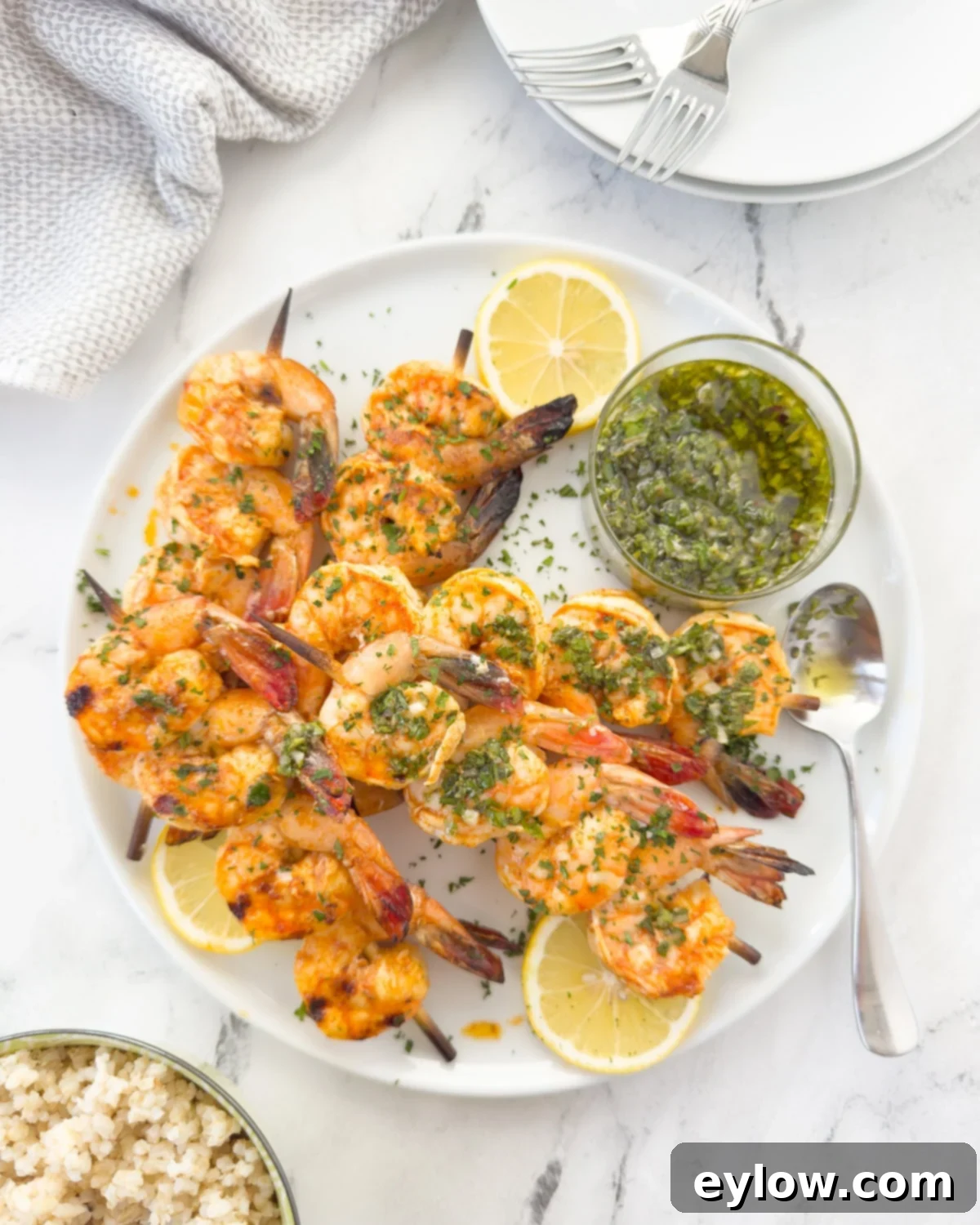 Grilled shrimp skewers piled on a round white plate with lemon slices and herb sauce.