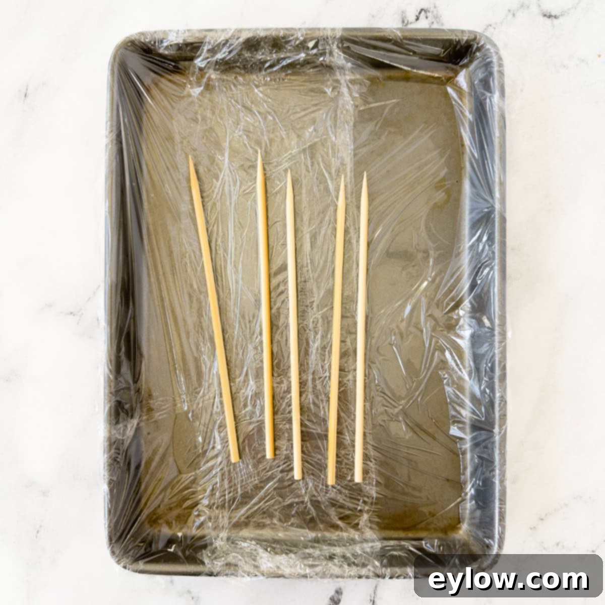 Bamboo skewers arranged on a plastic wrap lined sheet tray. 
