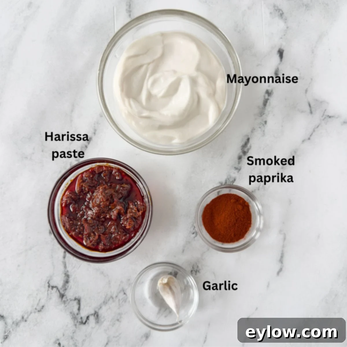 Harissa aioli ingredients in glass prep bowls on a marble counter.
