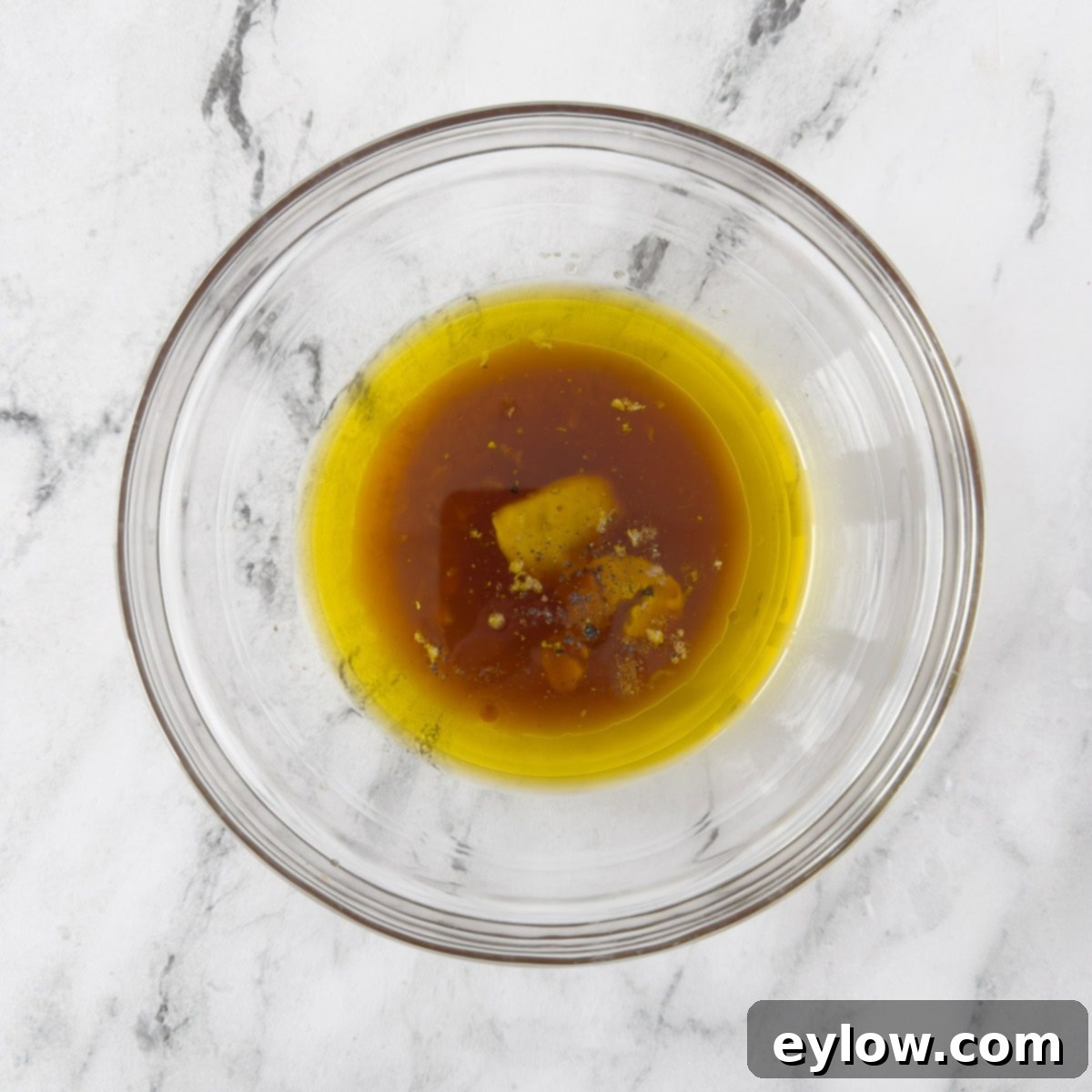 All the essential ingredients for a French vinaigrette, including oil, vinegar, Dijon mustard, and garlic, are combined in a clear glass bowl, ready for whisking.
