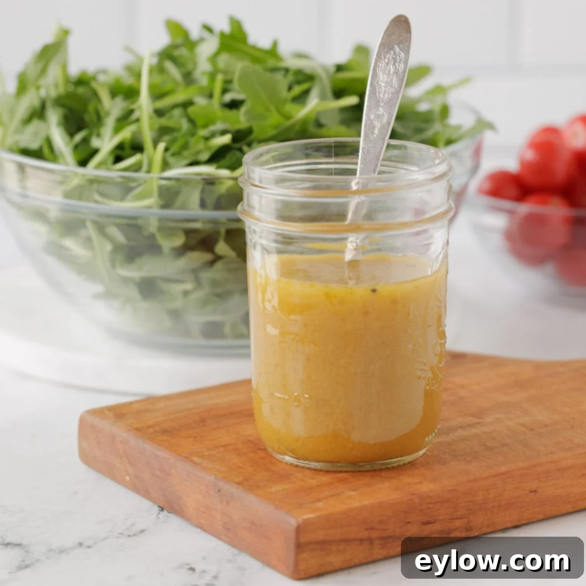 A small, elegant glass jar of homemade French vinaigrette sits on a clean kitchen counter, with a blurred background of fresh salad greens, emphasizing its readiness for a healthy meal.