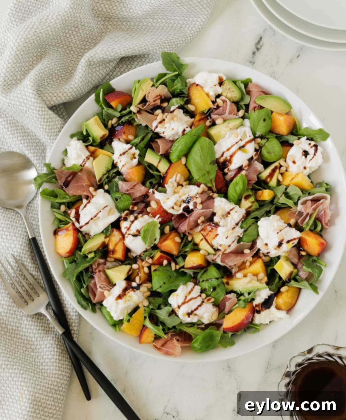 Burrata Peach and Prosciutto Summer Salad 2 A colorful burrata and peach salad with greens, avocado, basil, and nuts in a white salad bowl.