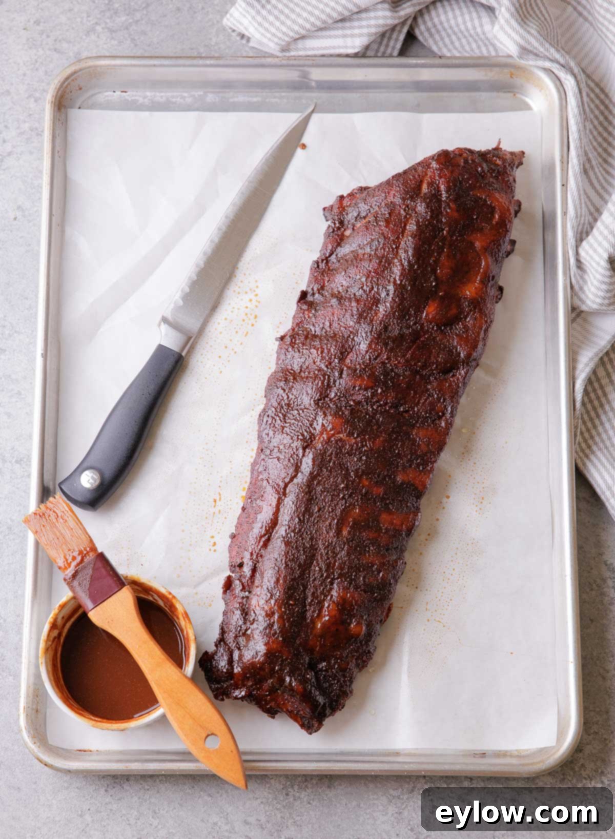 A rack of smoked baby back ribs before slicing, with a knife and sauce on a sheet tray.