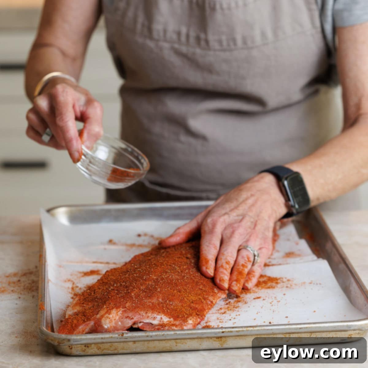 Seasoning baby back ribs with a bright red dry rub on a sheet pan.