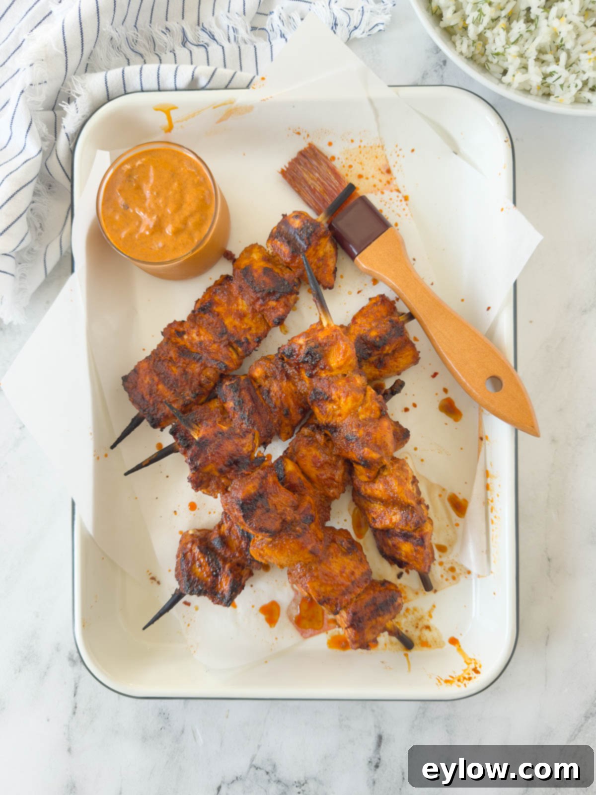 Fiery Harissa Chicken Skewers 2 Vibrantly orange-red harissa chicken skewers arranged on a tray, ready for grilling, with a pastry brush and a bowl of harissa sauce beside them.