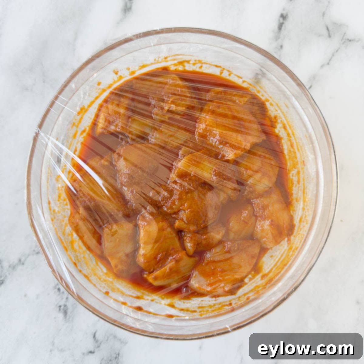 Fiery Harissa Chicken Skewers 6 A bowl of chicken marinating in harissa sauce, covered with plastic wrap, resting in the refrigerator.