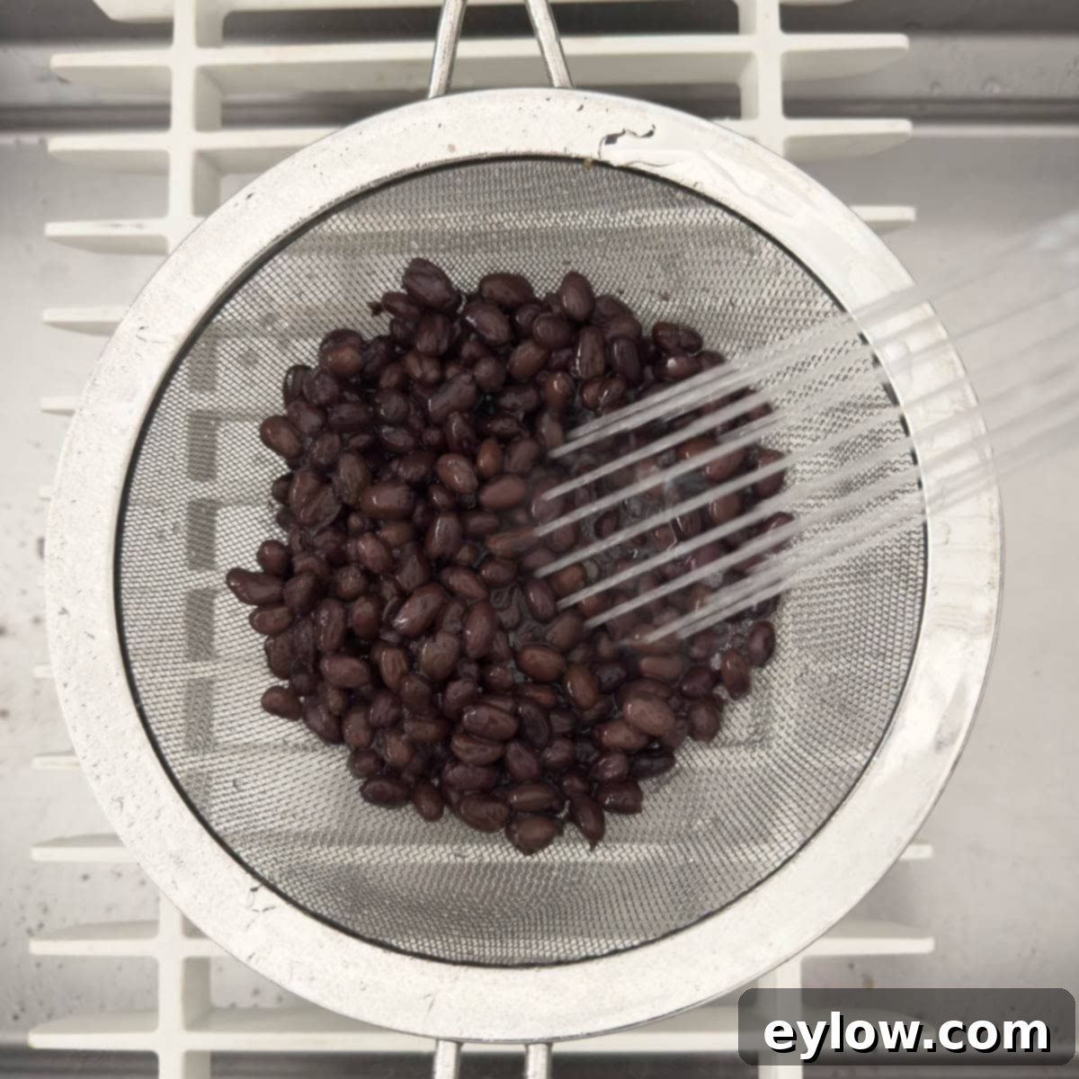 Black beans being thoroughly rinsed and drained in a sieve under running water from a sprayer in a kitchen sink.