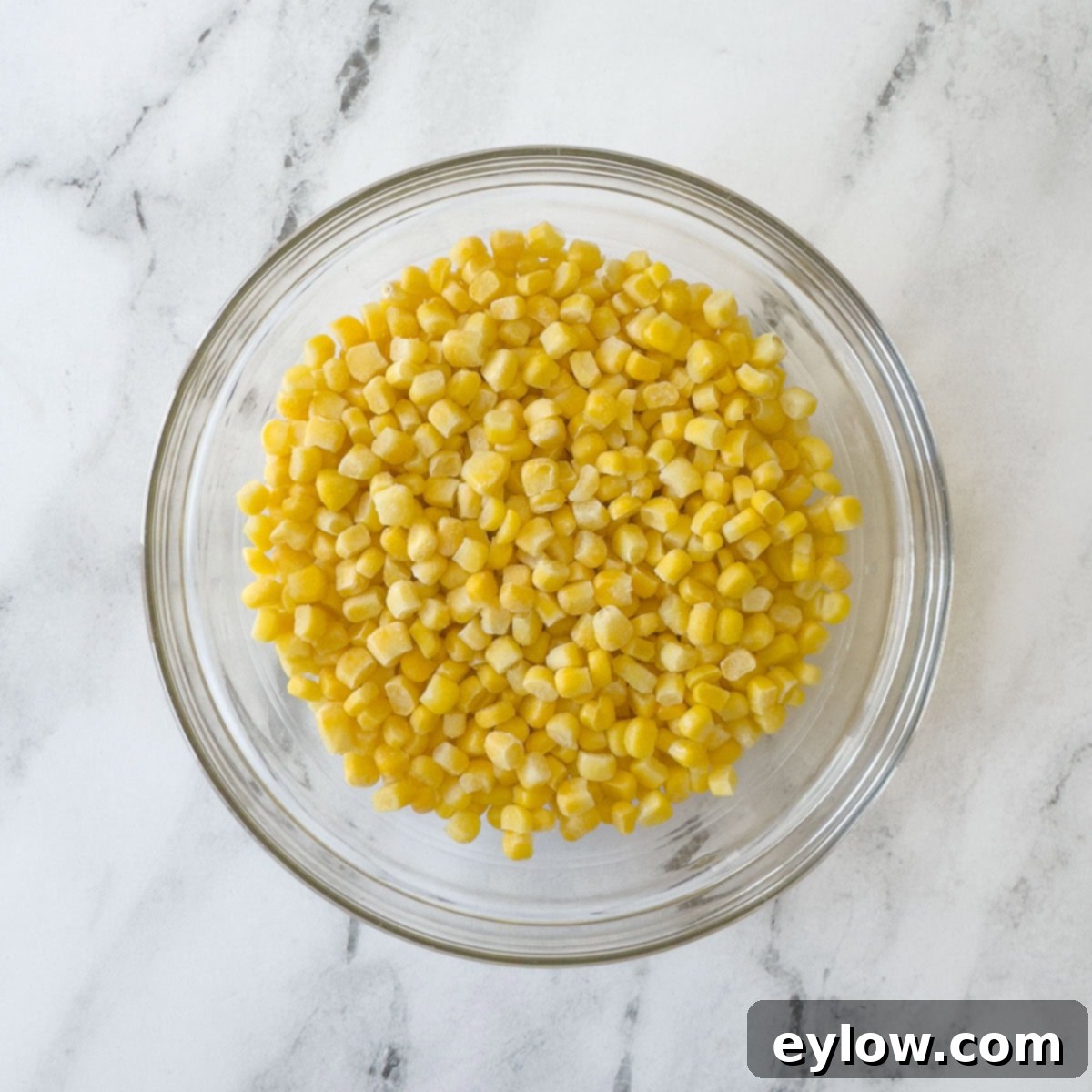 A sparkling glass bowl filled with golden yellow corn kernels rests on a elegant marble kitchen counter, showcasing vibrant freshness.