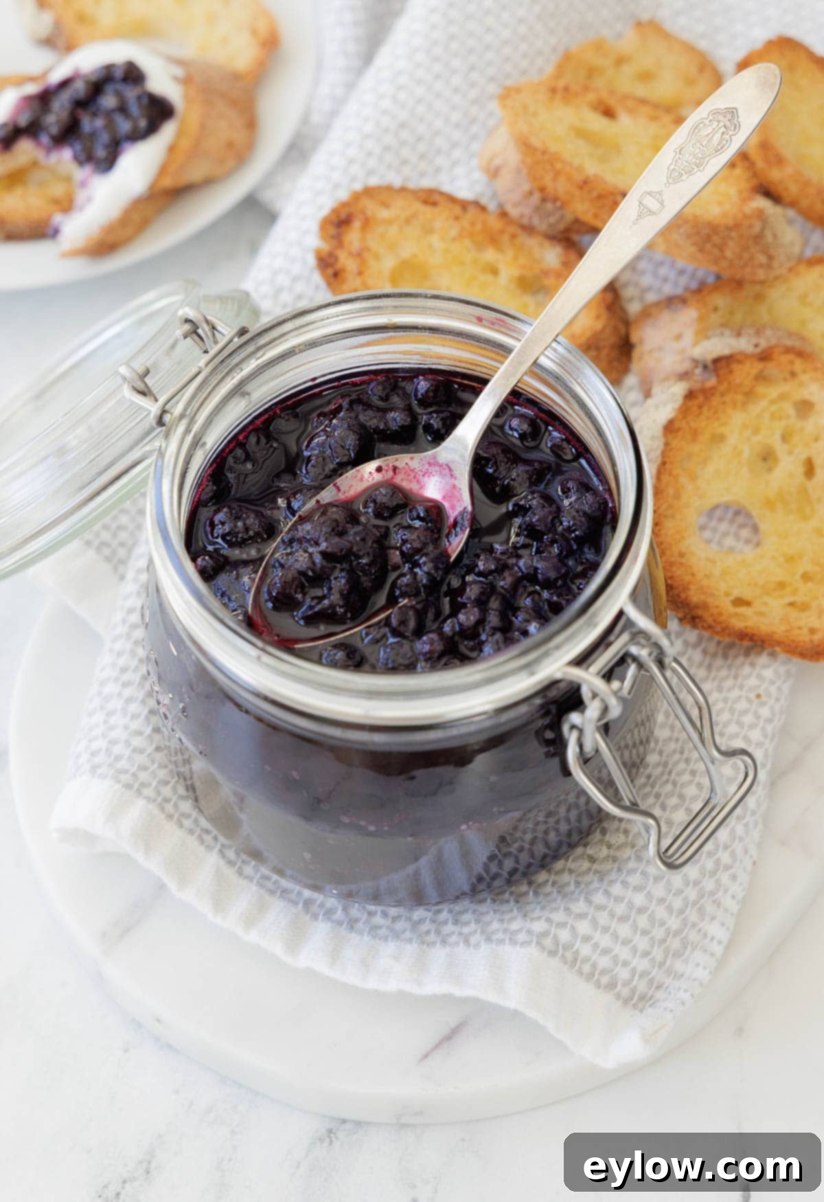 Effortless Blueberry Compote 2 A charming glass jar brimming with luscious, deep purple blueberry compote. A silver spoon rests beside it, ready to scoop, accompanied by golden, crisp crostini for a perfect pairing.