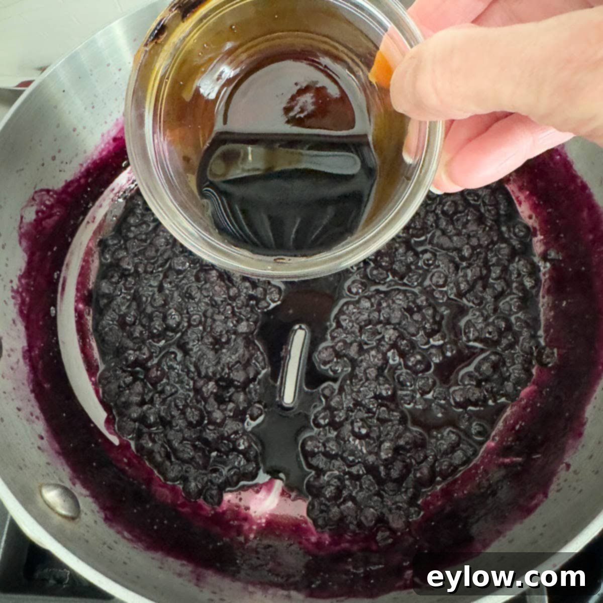 Effortless Blueberry Compote 7 A hand is seen pouring a stream of dark balsamic vinegar into a simmering pan of blueberries, enhancing the compote's flavor profile.
