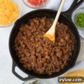 Taco meat in a cast iron pan with various fresh taco fixings like cheese, cilantro, and lime around it.