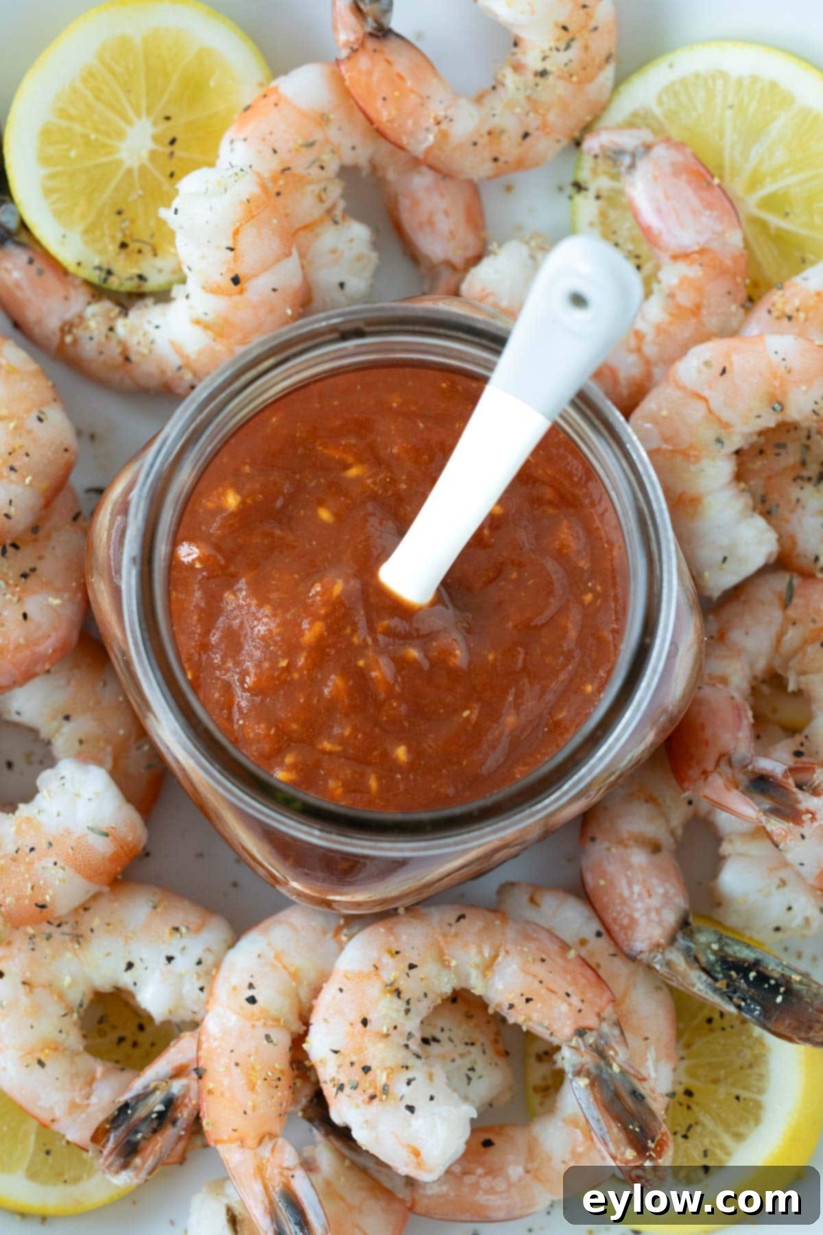 A glass jar of vibrant red homemade cocktail sauce with a white spoon, surrounded by cooked shrimp and fresh lemon slices on a white background.