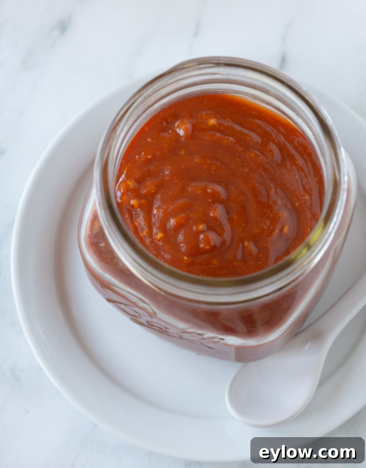 A jar of bright red homemade cocktail sauce (seafood sauce) with a white spoon on a white plate.