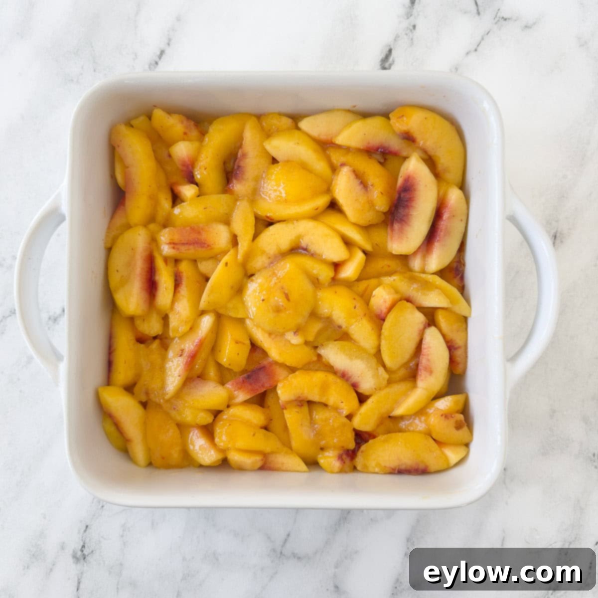 A square baking dish filled with sliced peaches to make ap each cobbler. 