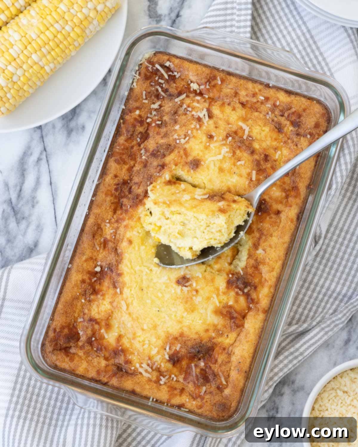 A golden corn souffle casserole in a glass baking dish with corn cobs and parmesan nearby.