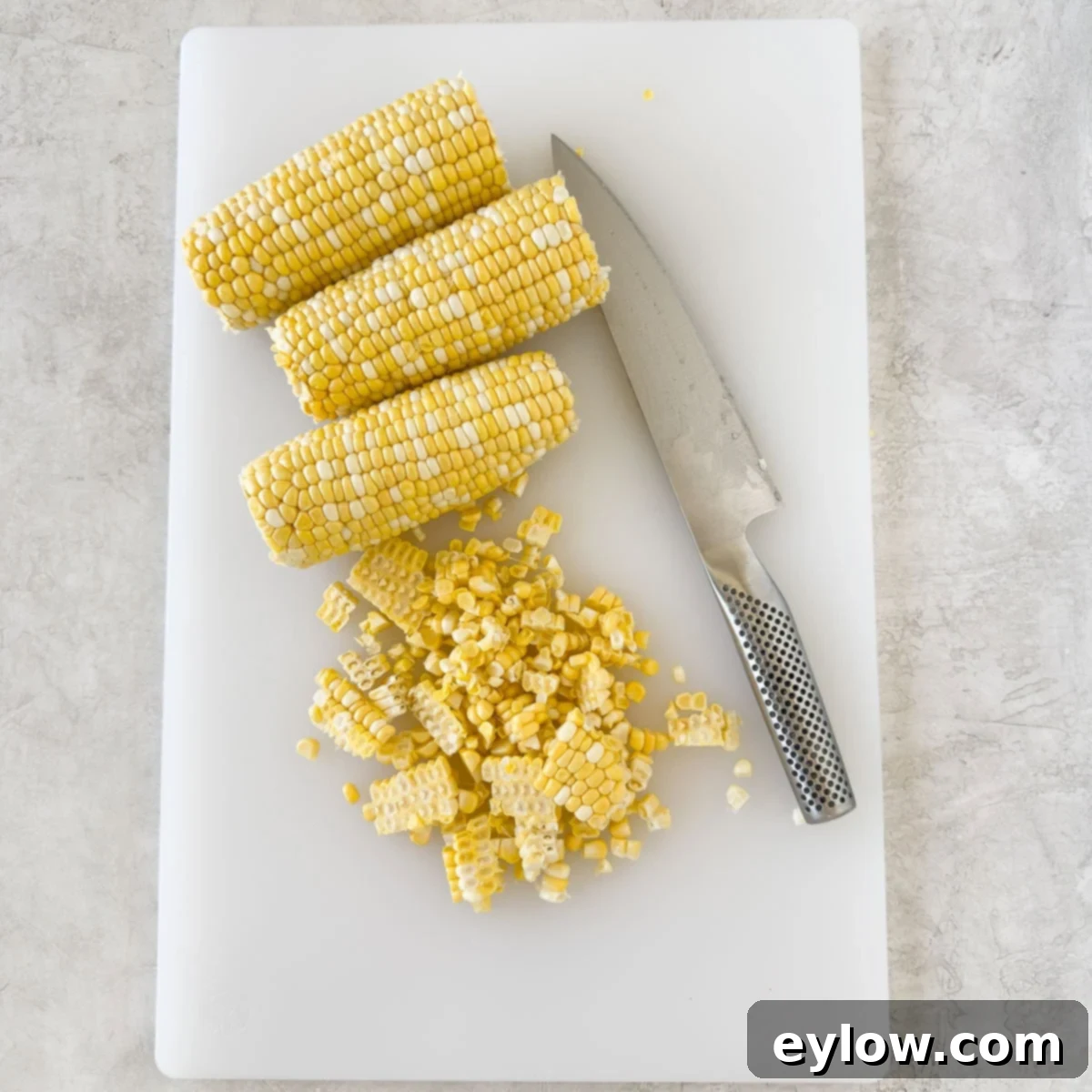 Cutting corn kernels off the cob on a white cutting board.