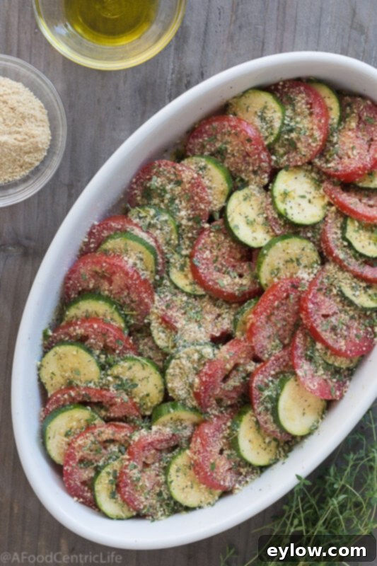 tomato zucchini casserole | AFoodCentricLife.com Layers of zucchini and tomato slices arranged in an oval baking dish with a cheese topping, ready for baking.