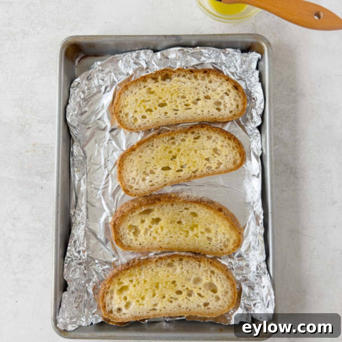 Slices of French bread brushed with a mixture of garlic oil and butter, arranged on a sheet tray for toasting.