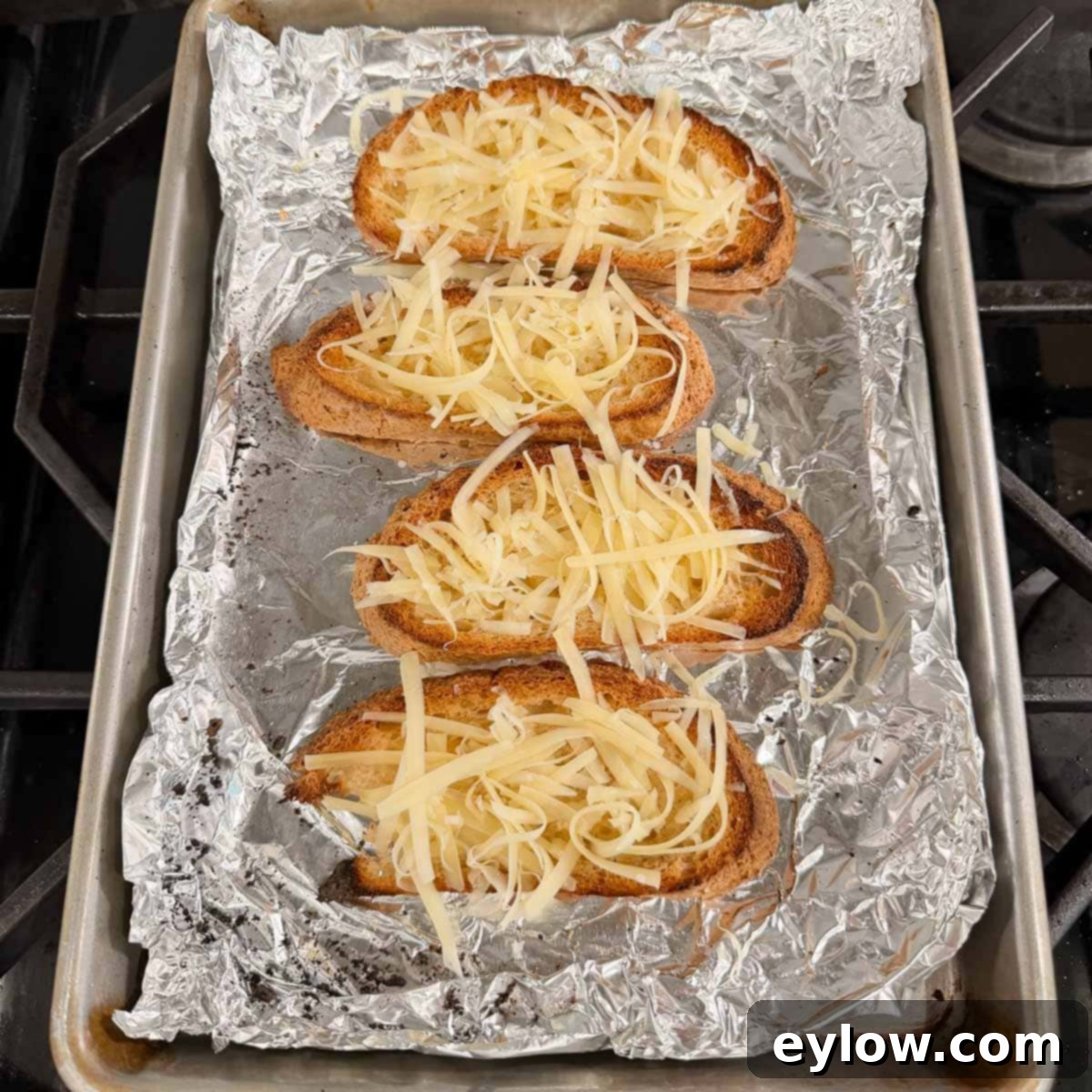 Garlic cheese toast, topped with grated Gruyere, ready for broiling on a foil-lined baking tray.