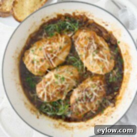 Four golden chicken breasts topped with caramelized onions, fresh herbs, and melting Gruyère cheese, in a shallow pan.