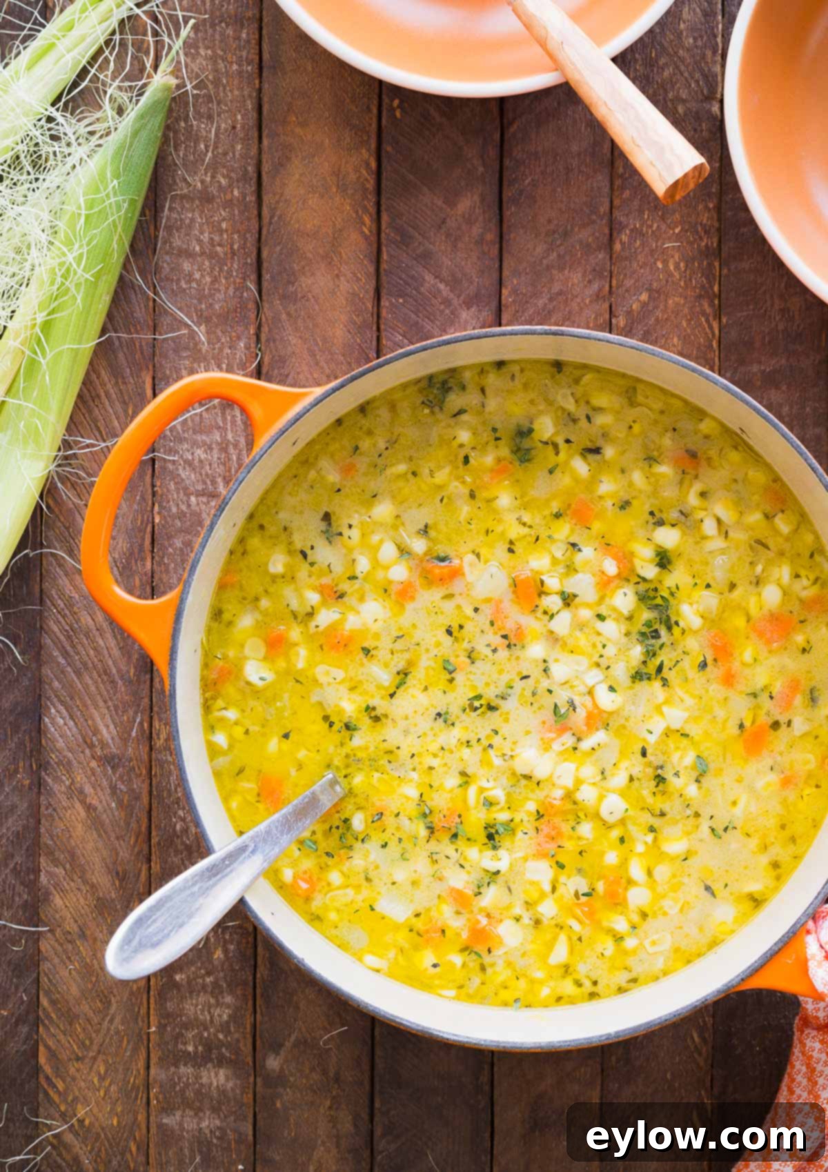 Creamy Dairy-Free Potato Corn Chowder 2 An orange dutch oven filled with creamy yellow corn chowder on a wooden counter, garnished with fresh herbs.