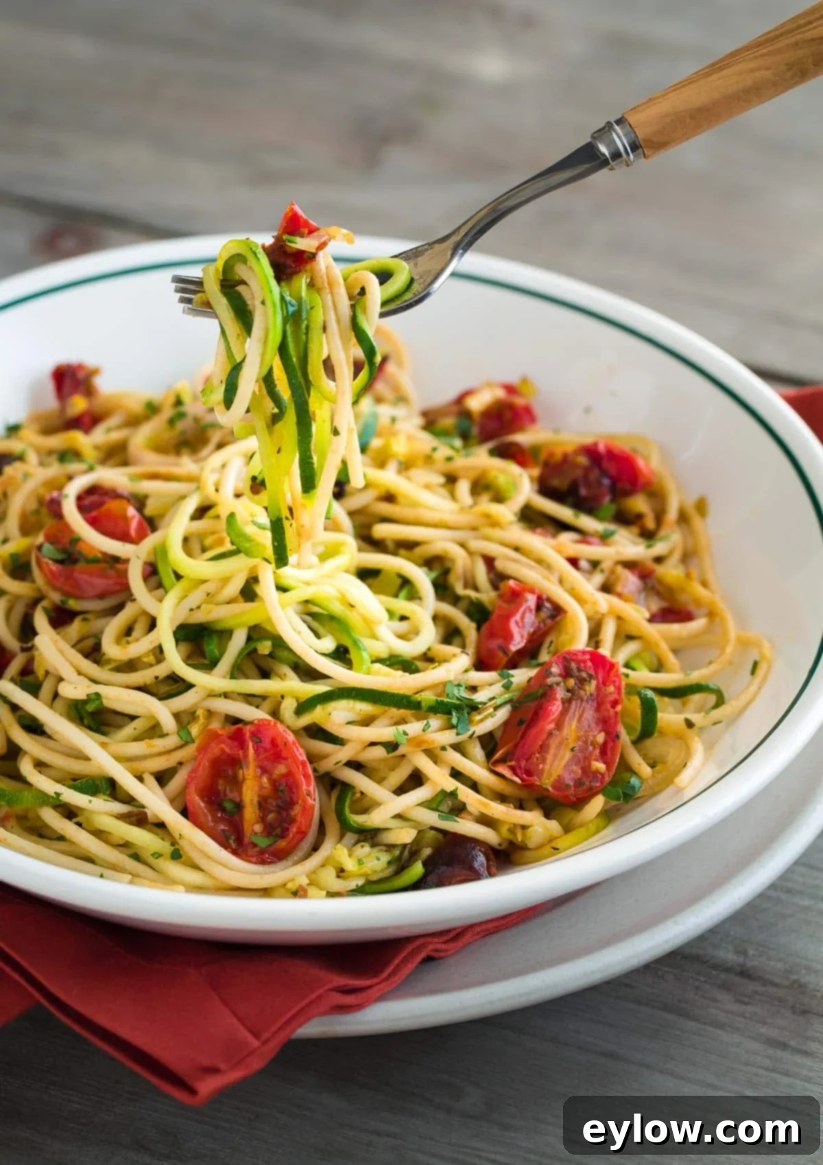 A bowl of zucchini noodles and pasta noodles with roasted red tomatoes on a fork.
