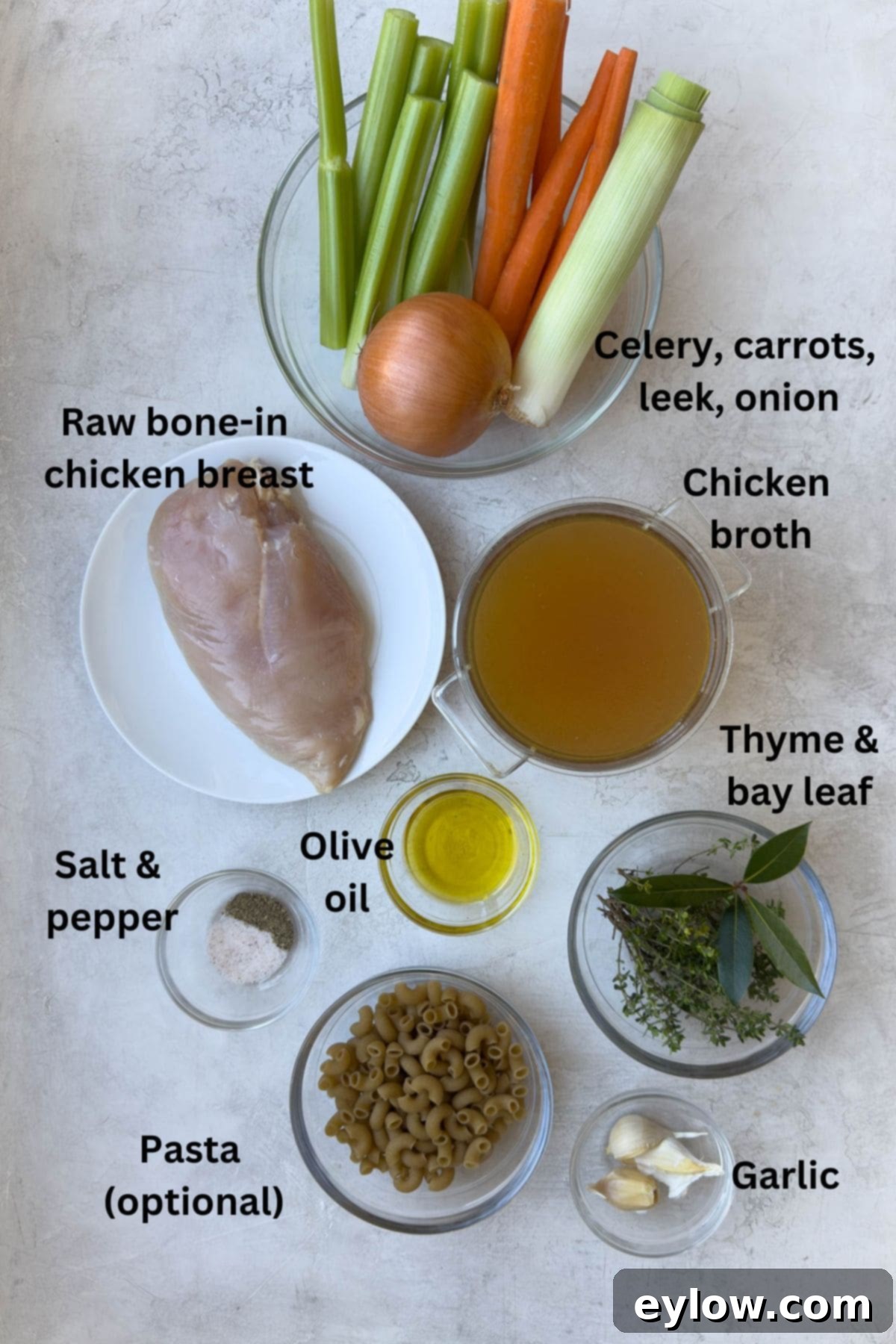 A flat lay photograph of fresh ingredients for chicken vegetable soup laid out on a wooden cutting board, including carrots, celery, onion, leeks, garlic, thyme, parsley, bay leaf, and raw chicken breast.
