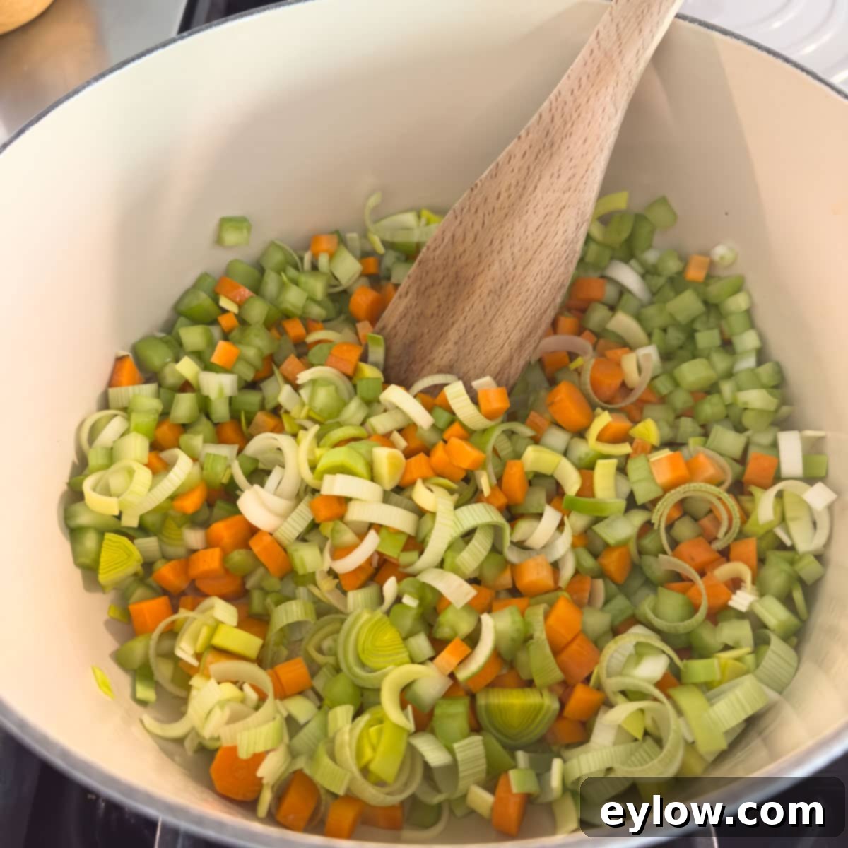 A pot on a stovetop with chopped onions, celery, carrots, and leeks beginning to sauté in olive oil.