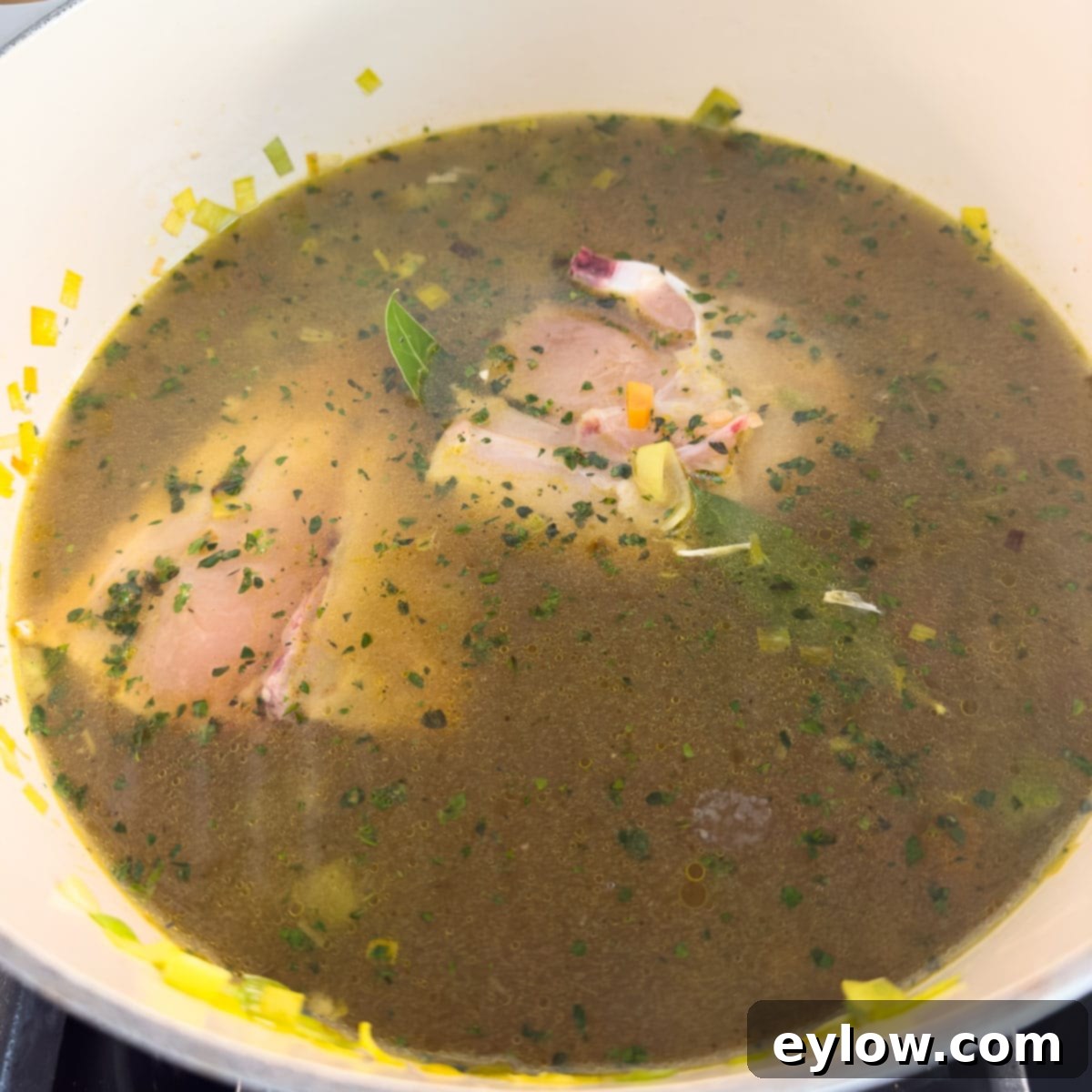 A raw, bone-in chicken breast submerged in a pot of simmering chicken broth with sautéed vegetables.