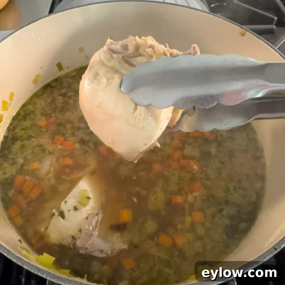 A cooked chicken breast being carefully removed from a pot of simmering soup using kitchen tongs.