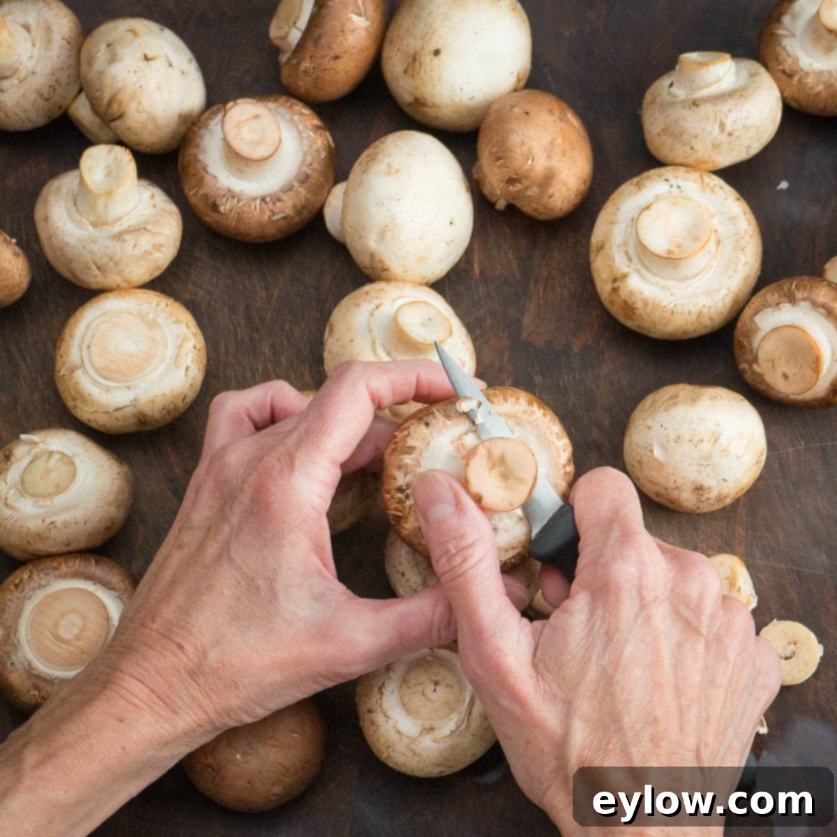 Hands trimming mushroom stems with a paring knife on a walnut cutting board.
