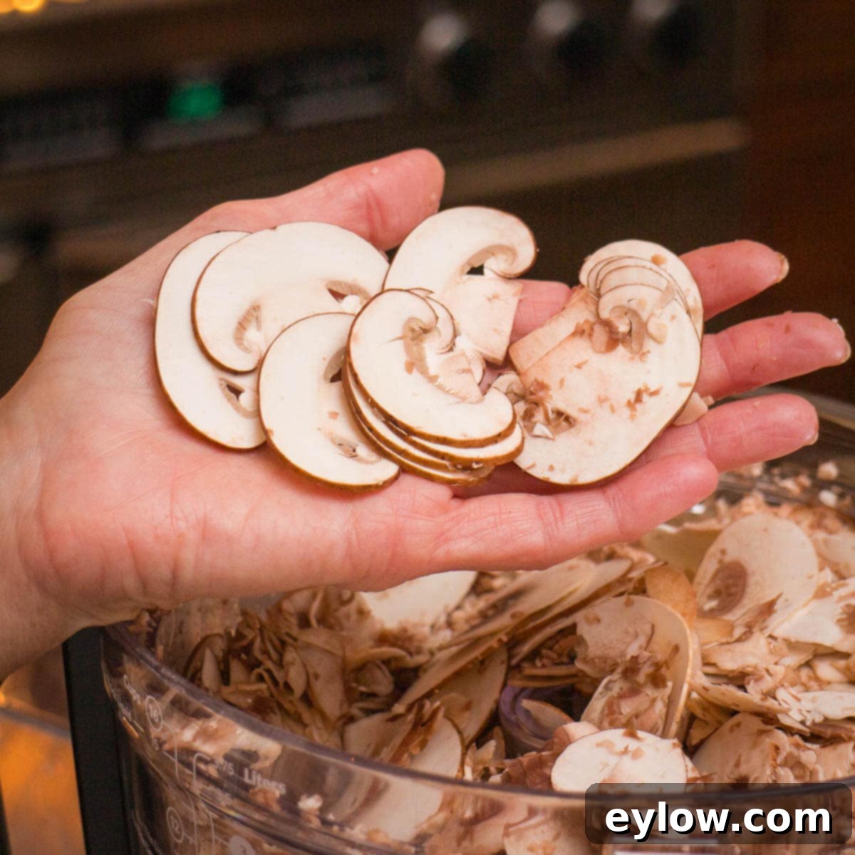 Hands showing thinly sliced mushrooms above the bowl of a food processor.