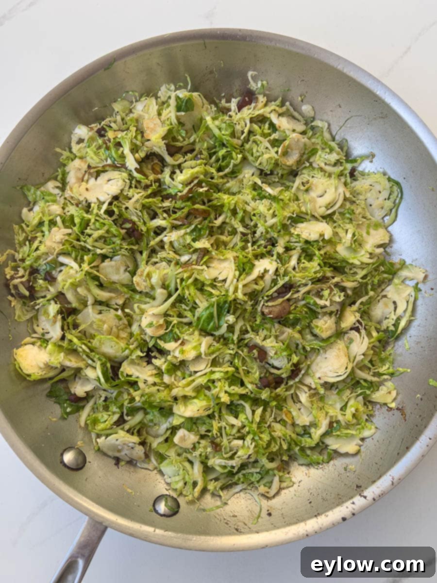 Beautifully cooked shaved Brussels sprouts, tender and vibrant, are tossed with crisp bacon bits in a stainless steel sauté pan, indicating the dish is complete and ready to be served and enjoyed.