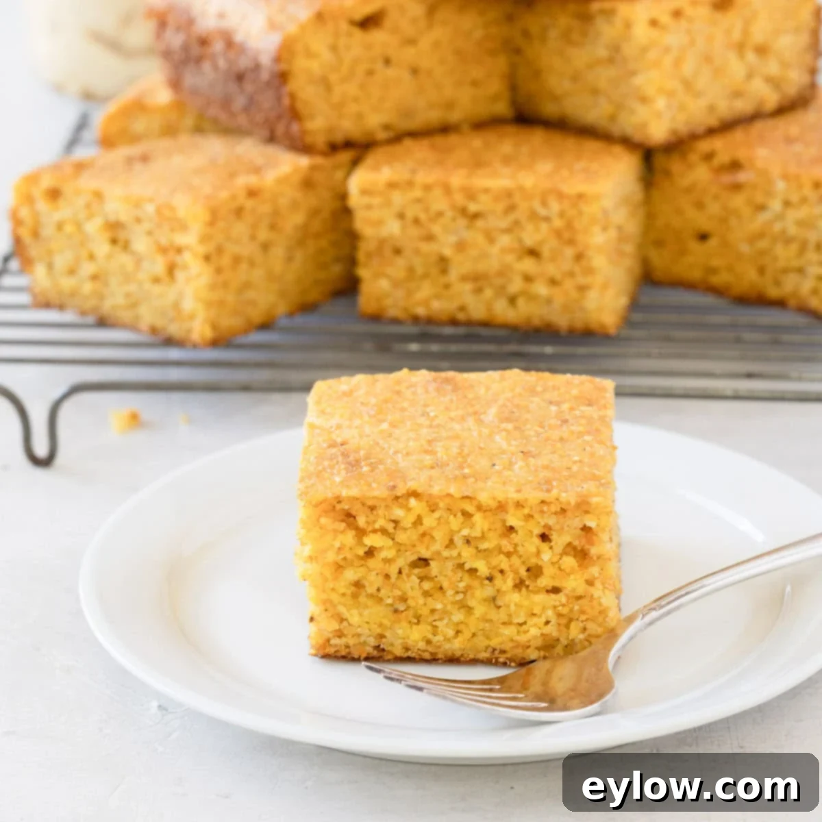 A single slice of moist, golden gluten-free cornbread rests on a white plate with a silver fork, with more slices stacked behind.