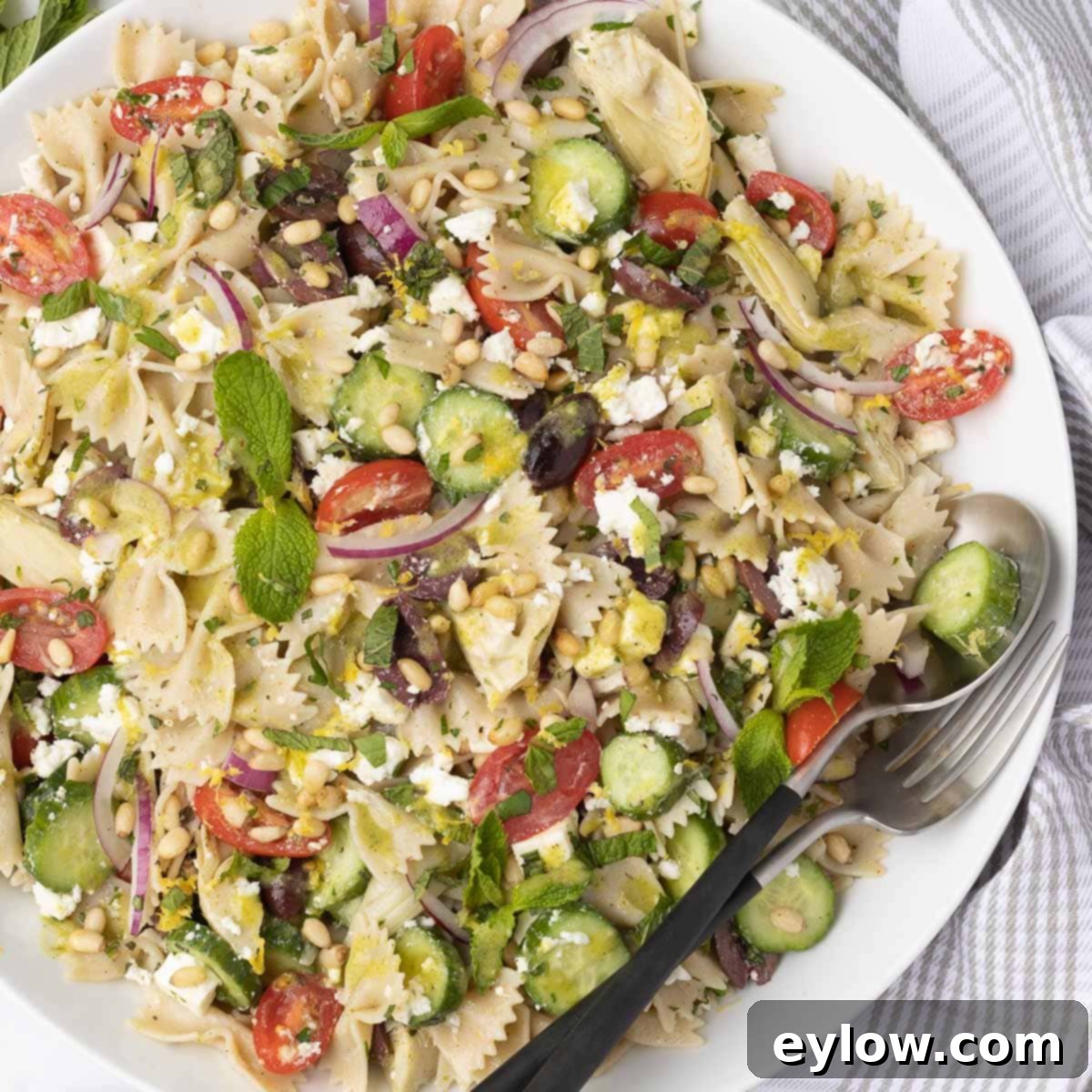 White bowl of colorful farfalle pasta salad with serving utensils.