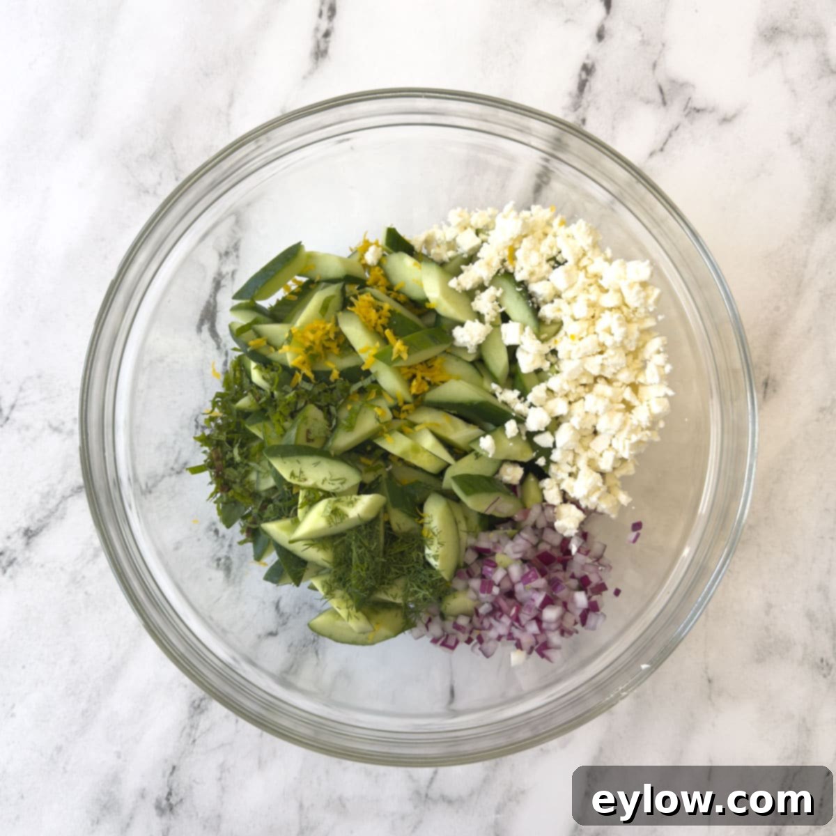 Crisp Cucumber Dill Feta Medley 8 A glass bowl with chopped cucumber, red onion, feta cheese, herbs, and lemon zest.