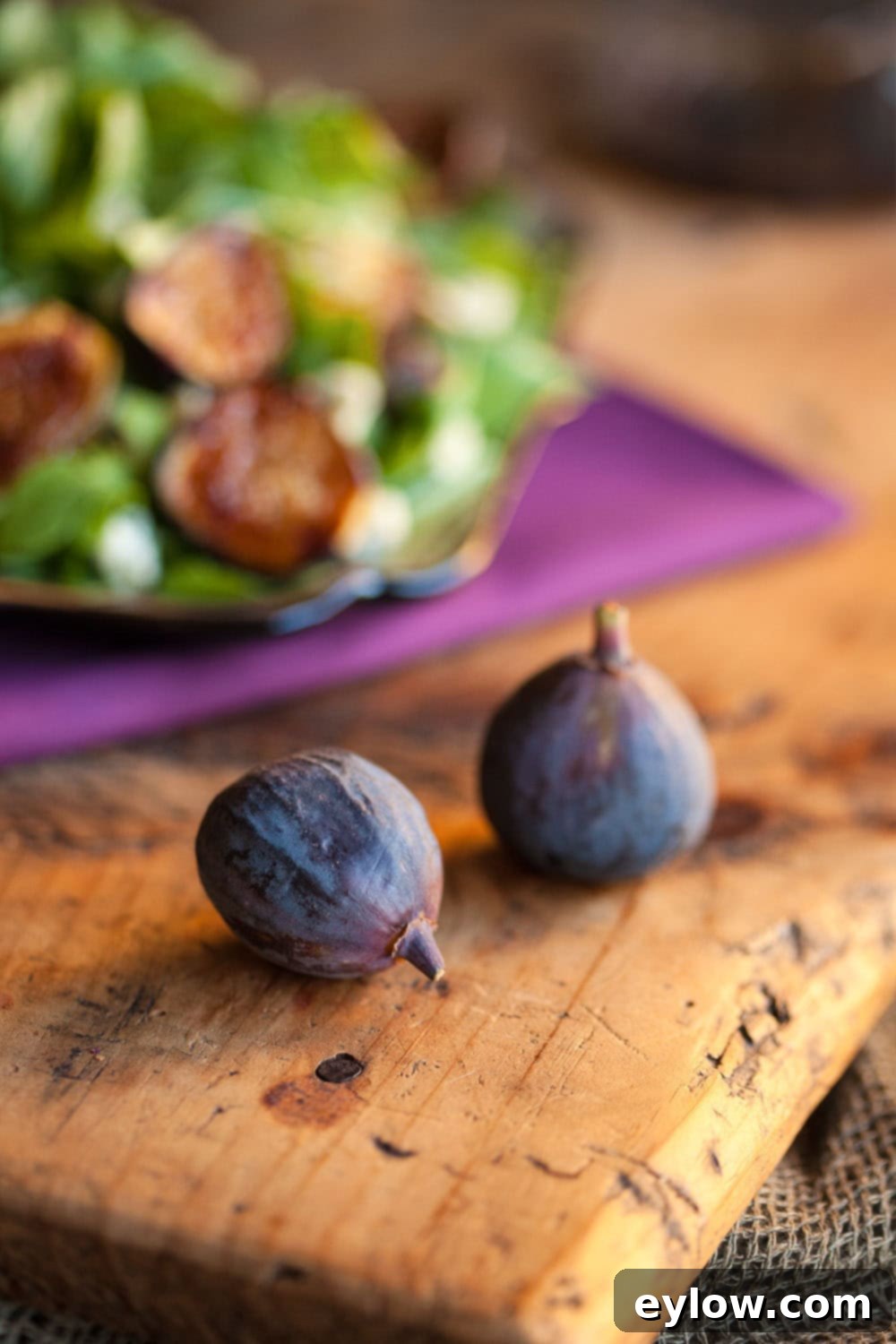 Two dark Black Mission figs on a weathered wooden cutting board, with fresh salad greens blurred in the background, suggesting preparation for a delicious fig salad.