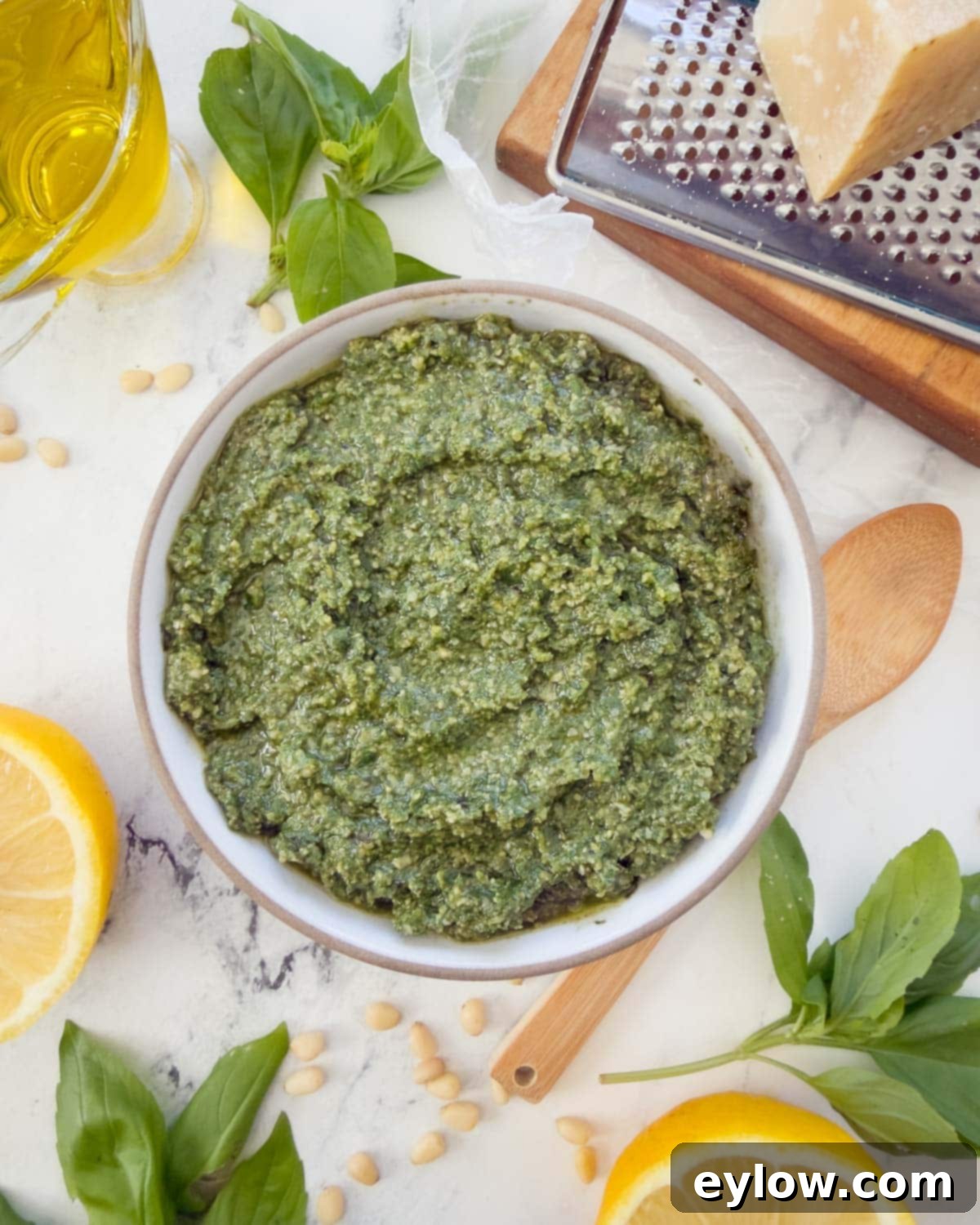 Genovese Basil Pesto: The Original Italian Recipe 2 A bowl of bright green Pesto Genovese, or classic basil pesto, surrounded by fresh basil leaves, olive oil, lemon wedges, and a block of Parmesan cheese, signifying its fresh, aromatic ingredients.