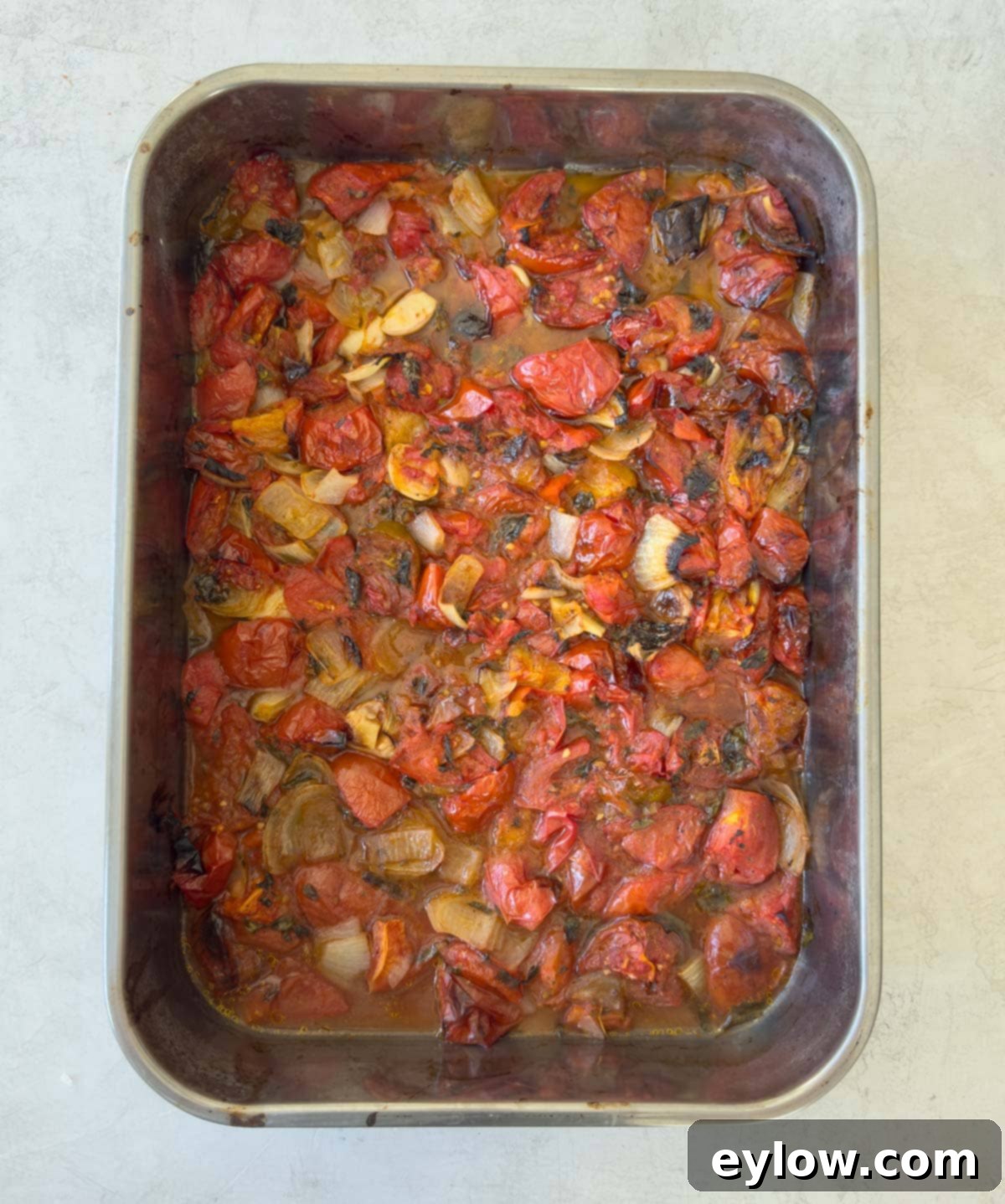 A stainless steel roasting pan of roasted tomatoes, onions, herbs, and garlic.