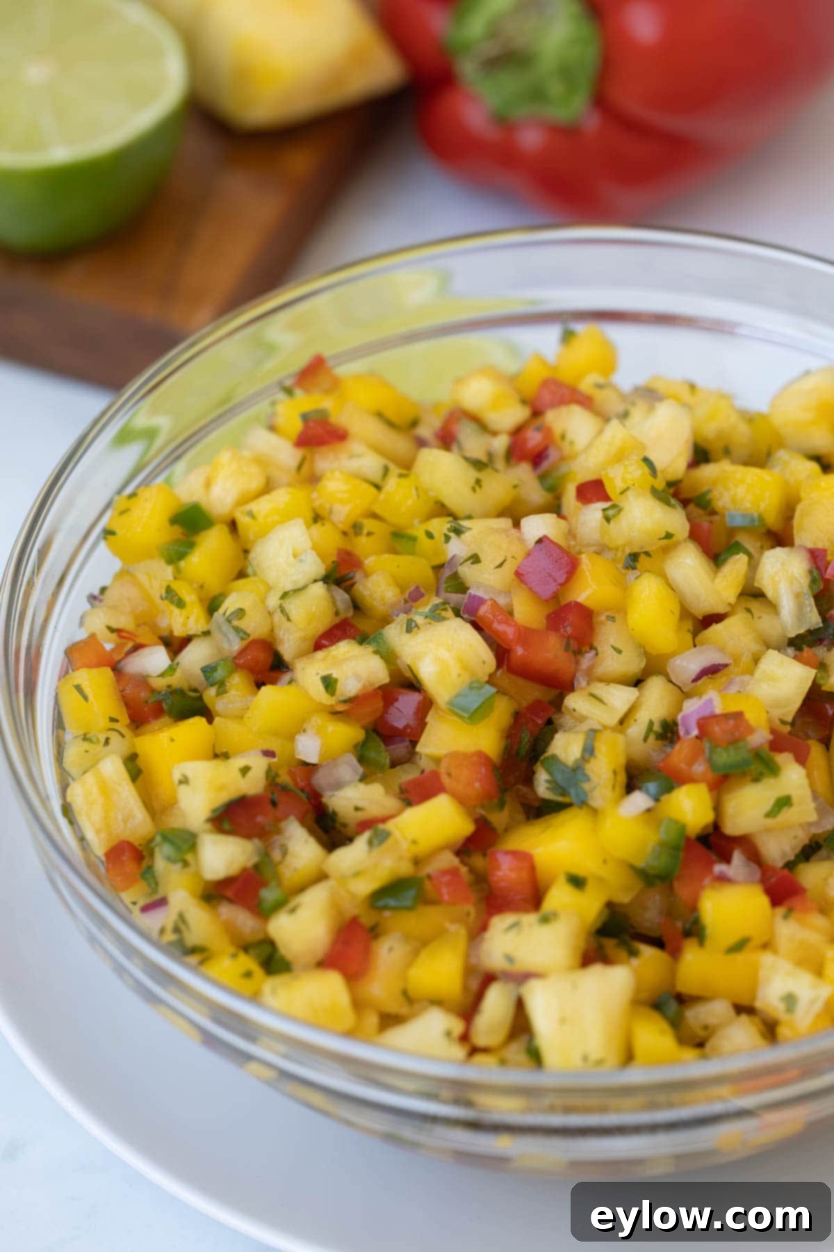 A glistening glass bowl overflowing with tropical fruit salsa, featuring chunks of pineapple, mango, vibrant red and green peppers, and fresh green herbs.