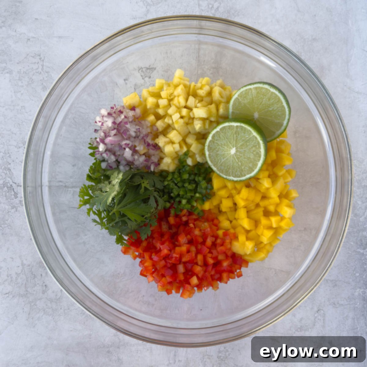 A large glass bowl showcasing a vibrant mix of diced pineapple, mango, red and green peppers, red onion, and fresh herbs, ready to be tossed into salsa.
