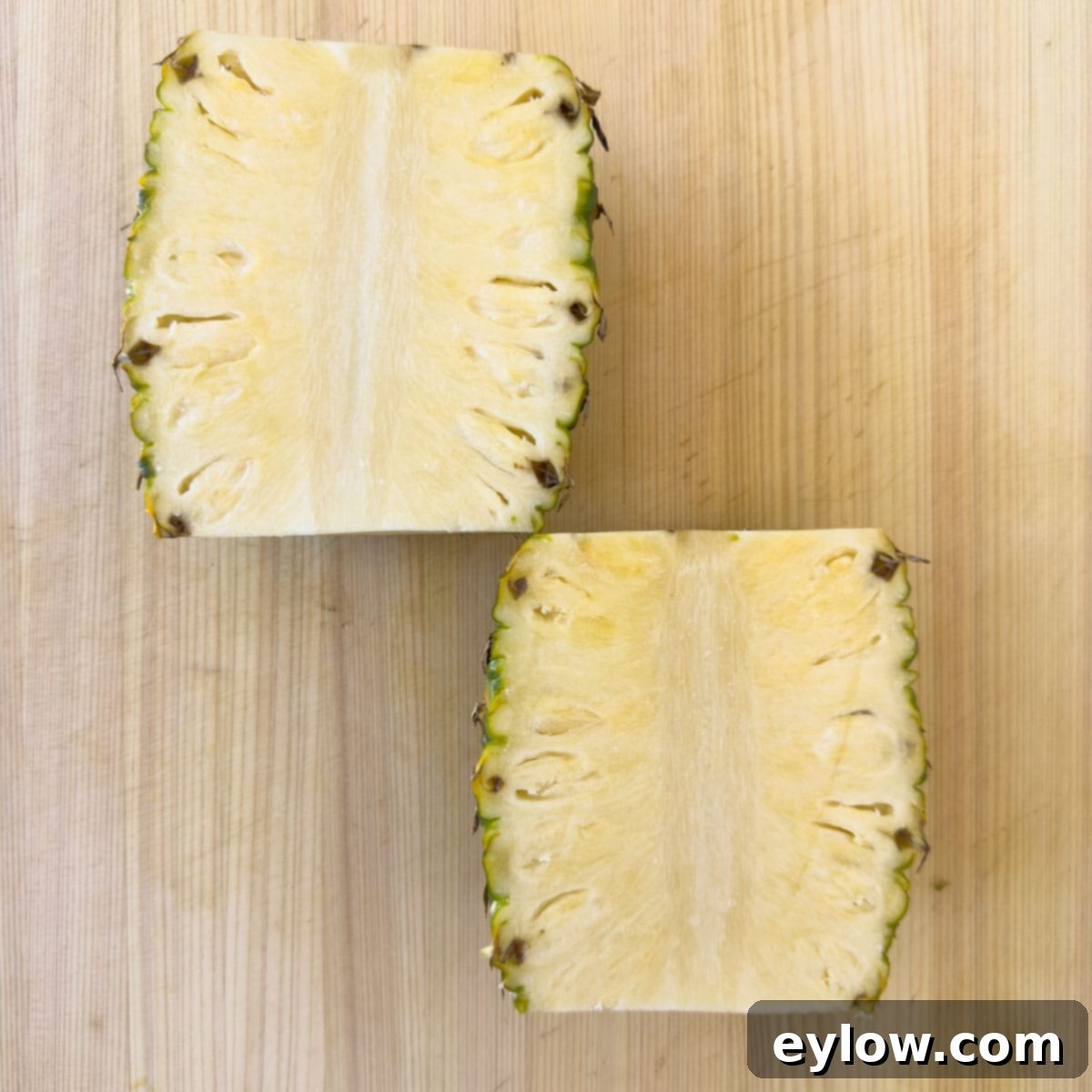 A pineapple, with its top and bottom removed, standing upright on a cutting board, being cut in half vertically.