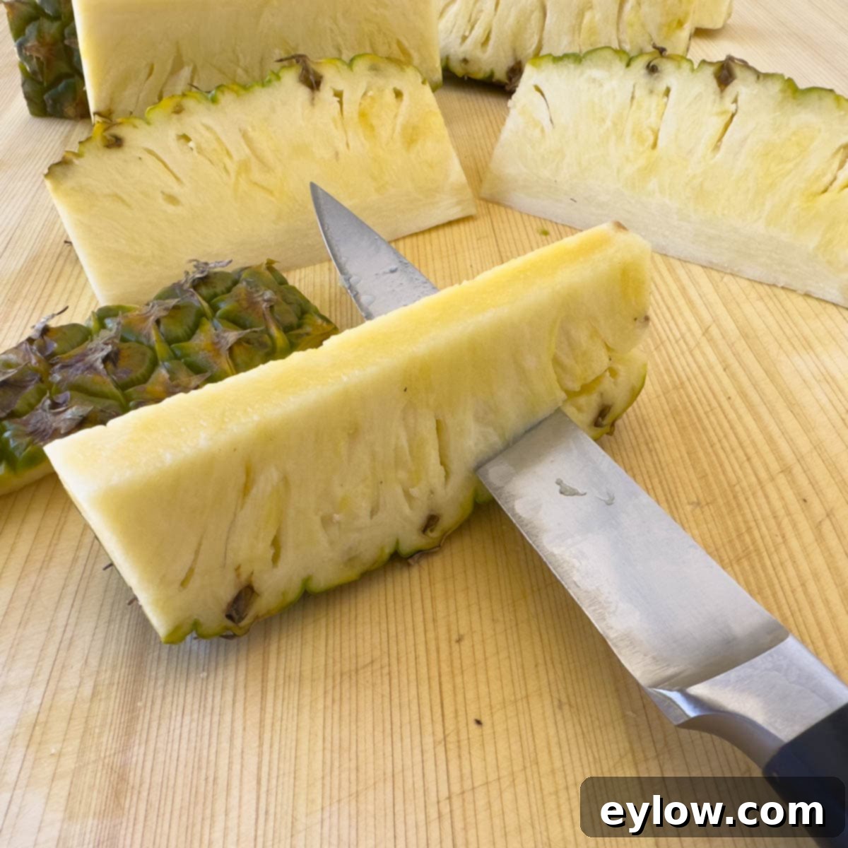 A pineapple spear on a cutting board, being de-skinned with a knife, showing the removal of the prickly exterior.