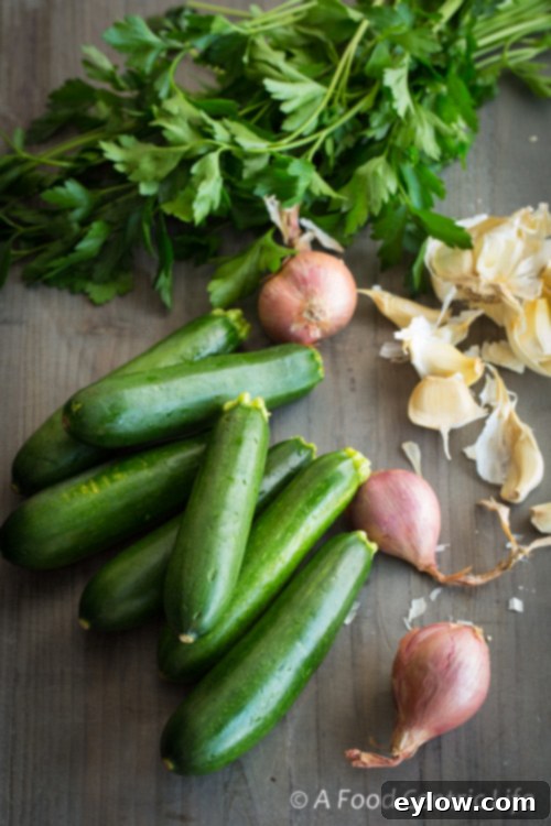 Golden Parmesan Zucchini Gratin 3 A vibrant selection of fresh ingredients laid out on a counter, including green zucchini, a bunch of fresh parsley, individual garlic cloves, and several shallots, ready for making zucchini gratin.